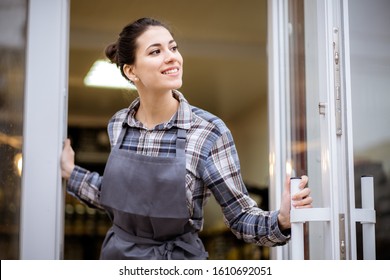 Mixed Race Woman Worker Or Store Owner With Standing In The Doorway Of Her Grocery Shop Looking Aside Smiling. Portrait Of Girl Assistant Wearing Apron Welcoming Inviting Open Door Of Store.