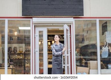 Mixed Race Woman Worker Or Store Owner With Standing In The Doorway Of Her Grocery Shop Looking Aside Smiling. Portrait Of Girl Assistant Wearing Apron Welcoming Inviting Open Door Of Store.