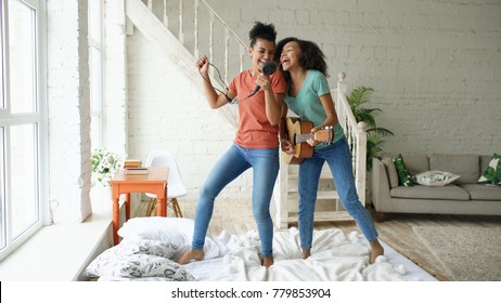 Mixed race young funny girls dance singing with hairdryer and playing acoustic guitar on a bed. Sisters having fun leisure in bedroom at home