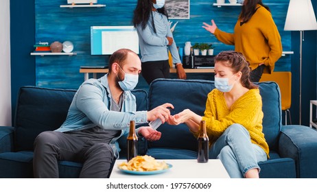 Mixed Races Friends With Medical Masks Disinfecting Hands With Sanitizer Gel Spending Time Together In Living Room. Diverse People Talking During Party, Enjoying Beer And Snacks In Global Pandemic