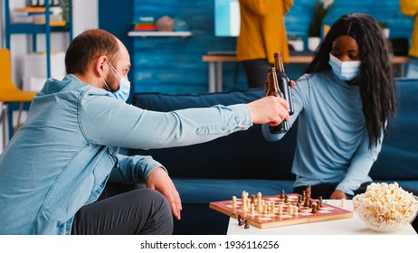 Mixed Races Of Friends Moving Pieces On The Chessboard Playing Board Games Wearing Face Masks Sitting On Couch In Living Room During New Normal Party. Keeping Social Distancing Against Global Pandemic