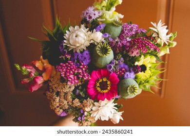 Mixed summer bouquet featuring poppy pods, strawflower, zinnias, and sunflowers with orange door background