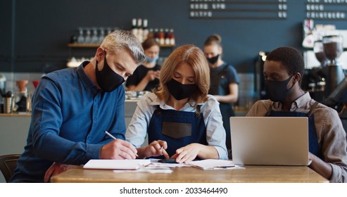 Mixed-race Cafe Waiters And Manager Discussing Working Schedule On Laptop. Restaurant Owner And Staff In Masks Looking At Computer Developing New Work Strategy During Quarantine Restrictions