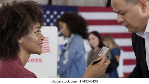 Mixed-race Staff Person Inspects Photo ID, Driving License, Presented By Hispanic Man At US Polling Station. Other Voters Soft Focus In Background With US Flag