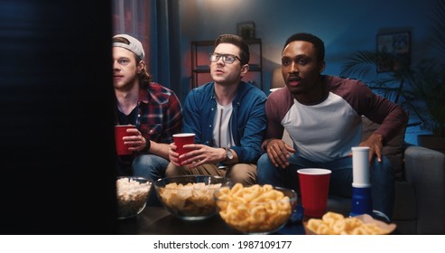 Mixed-race Young Handsome Males Sitting On Sofa In Living Room At Home At Night Watching Sport Channel On TV Feeling Sad Disappointed When Team Lose Game Match. Entertainment, Leisure Concept