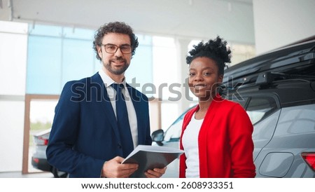Mixed-raced people glancing at camera at same time. Attractive man in business costume holding folder and standing next to pretty woman. Consultant and client happy about successful deal.