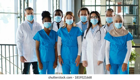 Mixed-races busy team of male and female doctors posing to camera and crossing hands in hospital. International group of medics in medical masks. Protected multi ethnic physicians and nurses in clinic