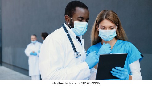 Mixed-races couple, man and woman, doctors in medical masks talking and using tablet device. Multi ethnic male and female physicians tapping and scrolling on gadget computer. Coworking in hospital.
