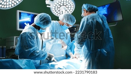 Mixed-races doctors during surgery in operating room. Professional surgeons in medical uniform and masks performing operation on patient under the bright lamp at hospital. Clinic concept