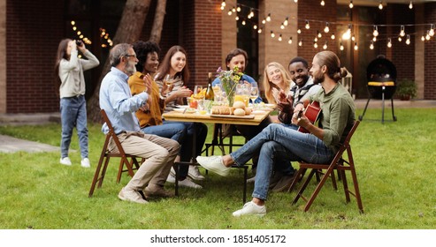 Mixed-races happy big family sitting at table with dinner at back yard outdoors and having fun. Guy playing on guitar. Musician entertaining at picnic. - Powered by Shutterstock