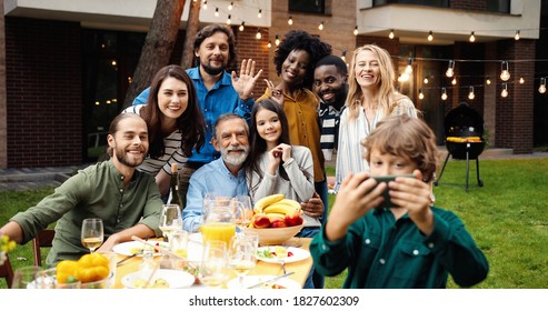 Mixed-races Happy Family At Party Dinner Outdoor In Yard Smiling And Posing To Smartphone Camera While Small Boy Taking Selfie Photo. Multi Ethnic People Making Photos Together At Barbeque Celebration