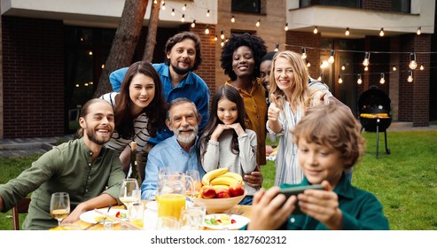 Mixed-races Happy Family At Party Dinner Outdoor In Yard Smiling And Posing To Smartphone Camera While Small Boy Taking Selfie Photo. Multi Ethnic People Making Photos Together At Barbeque Celebration