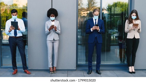 Mixed-races Male And Female People In Business Style And Medical Masks Standing With Gadgets And Keeping Social Distance. Coronavirus Pandemic