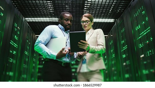 Mixed-races Man And Woman With Tablet Device Discussing Issue At Datacenter. Multiethnic Developers Standing At Servers Storage And Looking At Gadget. Male And Female Analytics Colleagues Talking.