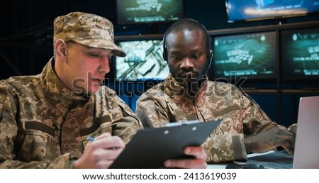 Mixed-races military male workers sitting in monitoring center and studying data from conflict zones. African American man in headset typing on laptop computer while Caucasian soldier reading document