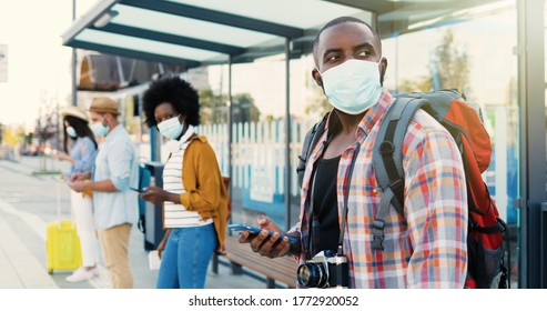 Mixed-races young males and females people im medical masks standing in line at bus stop. Keeping safe social distance. African American stylish man tourist outdoor waiting for transport. Tourists.