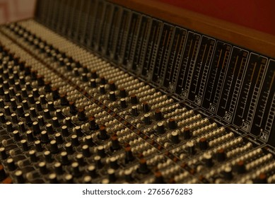 A mixing board is shown with many knobs and sliders in a studio setting. The equipment is lit up, indicating a busy recording session happening at night.