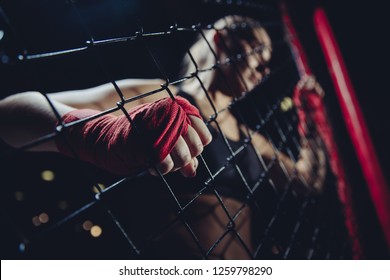 MMA is young girl in octagon during fights without rules gaining strength at grid of Boxing ring, hard clutching links fist wrapped red protective bandage. Selective focus