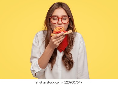 Mmm, So Delicious! Dark Haired Pretty Woman Eats Slice Of Italian Pizza, Keeps Eyes Closed From Pleasure, Enjoys Nice Taste, Wears Glasses And Shirt, Isolated Over Yellow Background. Eating Concept