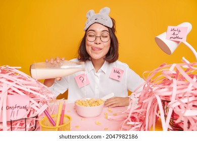 Mmm how delicious. Asian woman pours milk into cereals going to have breakfast licks lips dressed in white shirt and sleepmask poses at desktop isolated over yellow background. Work from home - Powered by Shutterstock