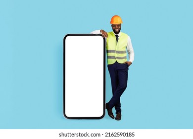 Mobile Ad. Handsome Black Civil Engineer Standing Near Big Blank Smartphone With White Screen, Smiling African American Builder Wearing Safety Vest And Hard Hat Recommending New Application, Mockup