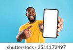 Mobile advertisement. Overjoyed black man pointing at cellphone with empty white screen on blue studio background, mockup for app or website. Cellphone display template