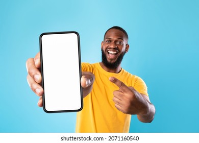 Mobile advertisement. Overjoyed black man pointing at cellphone with empty white screen on blue studio background, mockup for app or website. Cellphone display template