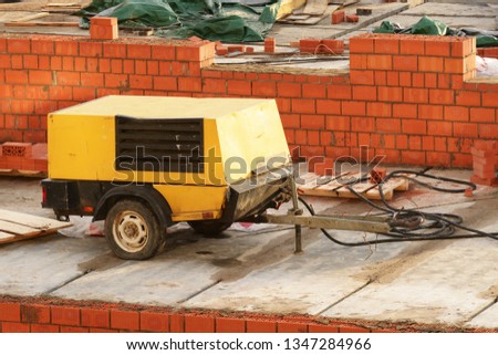 Mobile air compressor on construction site of a red brick house. It's device not new, painted black and yellow. It has pneumatic wheels and a hitch to attach to the car. 