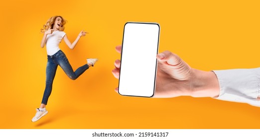 Mobile App Advert. Excited Cheerful Woman Jumping Up And Pointing Fingers At Huge White Empty Screen, Giant Hand Holding Big Cell Phone, Yellow Orange Studio Wall. Check This Gadget, Display Mock Up