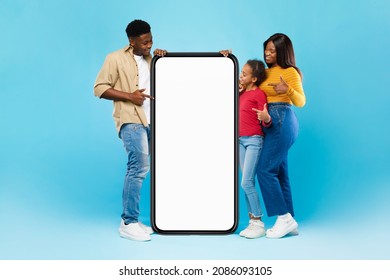 Mobile App Advert. Full Body Length Of Happy Black Family Leaning Pointing At Big Huge White Empty Smartphone Screen Standing At Blue Walll. Check This Website, Cellphone Display Mock Up, Free Space