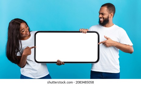 Mobile App Advertisement. Excited Black Couple Holding And Pointing At Big White Empty Smartphone Screen, People Presenting Cell Phone Display Mock Up On Blue Studio Background. Check This, Panorama