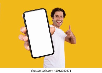 Mobile App Advertisement. Handsome Excited Man Showing Pointing At White Empty Smartphone Screen Posing Over Orange Studio Background, Smiling To Camera. Check This Out, Cellphone Display Mock Up