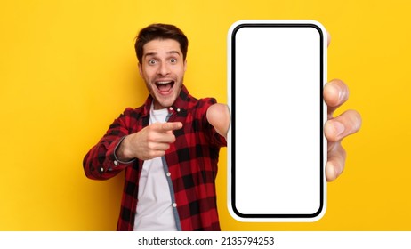 Mobile App Advertisement. Handsome Excited Guy Showing Pointing At White Empty Smartphone Screen Close To Camera Posing Over Yellow Studio Background. Check This Gadget Out, Cell Phone Display Mock Up