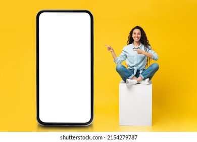 Mobile App Advertising. Happy Casual Lady Pointing Finger At Big Huge Blank Smartphone Display, Sitting On White Cube Presenting Huge Empty Cell Phone Screen, Yellow Wall, Mock Up, Full Body Length