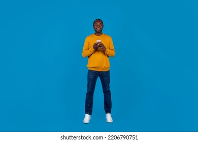 Mobile App Concept. Happy Handsome African American Mature Man In Stylish Casual Outfit Using Modern Cell Phone And Smiling On Blue Studio Background, Full Length Shot, Panorama With Copy Space