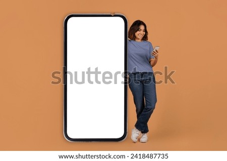 Mobile app. Middle eastern young woman stand next to big phone with white blank screen mockup copy space for advertisement, using smartphone, colorful background