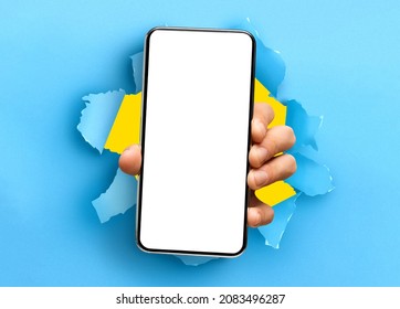 Mobile App Promo, Great Offer. Closeup Of Hand Holding Smartphone With White Empty Screen Showing Device Close To Camera Breaking Through Blue Paper Sheet. Blank Display With Free Copy Space, Mock Up