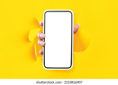 Mobile App Promo, Great Offer. Female Hand Holding Cell Phone With White Empty Screen Showing Device Close Up To Camera Breaking Through Yellow Paper Sheet. Blank Display With Free Copy Space, Mock Up