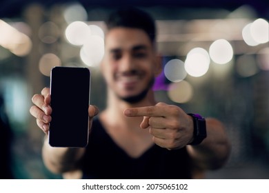 Mobile App. Smiling Arab Guy Pointing At Blank Smartphone With Black Screen, Middle Eastern Male Athlete Recommending New Fitness Application While Standing In Gym, Mockup Image With Copy Space