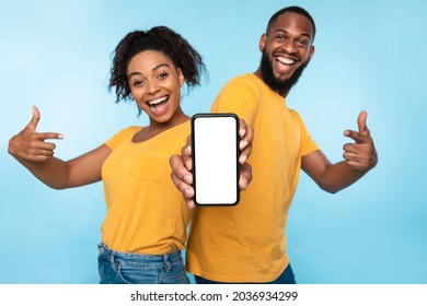 Mobile application ad. Excited Afro couple pointing at cellphone with blank screen, recommending new app on blue studio background, selective focus. Mockup for website