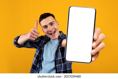 Mobile Application Ad. Excited Young Guy Showing Smartphone With Blank White Screen And Gesturing Thumb Up, Cheerful Man Recommending New App While Standing Over Yellow Studio Background, Mockup - Shutterstock ID 2057039480