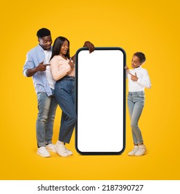 Mobile Application Ad. Full Body Length Of Young Black Family Pointing At Huge White Empty Smartphone Screen, Standing At Yellow Walll. Check This Website, Display Mockup, Copy Space