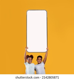 Mobile Application Ad. Happy Black Schoolchildren Hugging Holding Big Cell In Hands Above Head Showing Phone Empty Screen Recommending App On Yellow Orange Studio Background. Smartphone Display Mockup