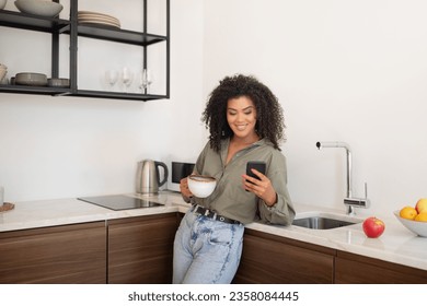 Mobile Application. Relaxed Black Woman Using App On Smartphone At Modern Kitchen Interior. Smiling Woman Holding Phone And Cup Of Coffee, Chatting In Social Media. Gadget And Communication - Powered by Shutterstock