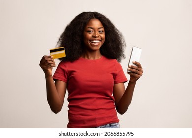 Mobile Banking. Happy Beautiful African American Woman Holding Smartphone And Credit Card, Cheerful Black Female Using Application For E-Commerce Or Shopping Online, Standing On White Background