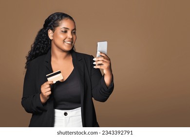 Mobile banking. Happy latin chubby lady using phone and holding credit card, making safe payment via bank app, brown studio background, free space. Business woman paying for service online - Powered by Shutterstock