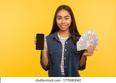 Mobile banking, online payment. Portrait of friendly happy girl in denim shirt holding dollar banknotes and cell phone with mock up blank display. indoor studio shot isolated on yellow background
