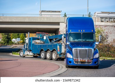 Mobile Big Rig Tow Truck With Lifting Boom And Three Driving Truck Rear Axles Towing Broken Blue Big Rig Semi Truck Tractor With Emergency Lights On The Grill Going Under The Bridge To Repair Shop 