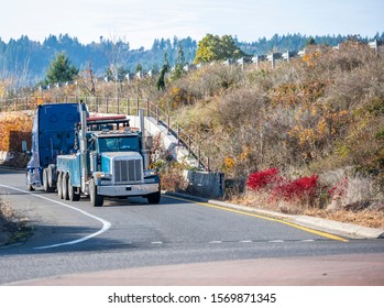 Mobile Big Rig Tow Truck With Lifting Boom And Three Driving Truck Rear Axles Towing Broken Blue Big Rig Semi Truck Tractor With Emergency Lights On The Grill Going Under The Bridge To Repair Shop 
