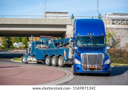 Mobile big rig tow truck with lifting boom and three driving truck rear axles towing broken blue big rig semi truck tractor with emergency lights on the grill going under the bridge to repair shop 